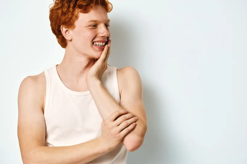 Young man with red hair wearing white tank top smiling and touching his face posing against white background for casual lifestyle and happiness concept.
