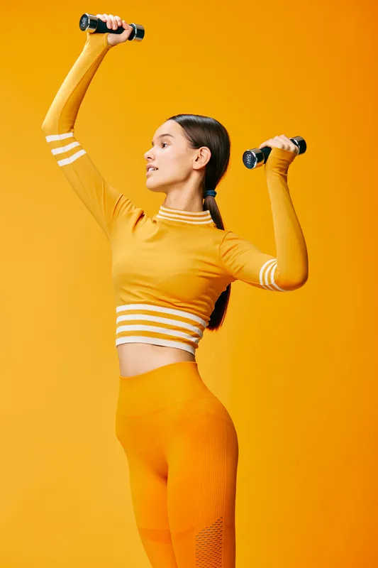 Young woman in vibrant orange workout outfit lifting small dumbbells in an energetic overhead exercise pose, studio fitness portrait with matching bold orange background.