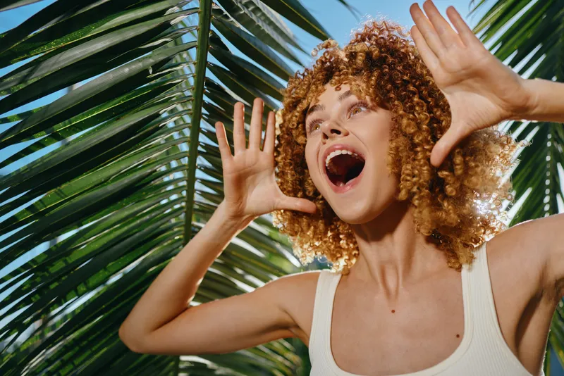 curly woman shouting joyfully near tropical palm leaves outdoors, expressing excitement and energy under natural sunlight in summer