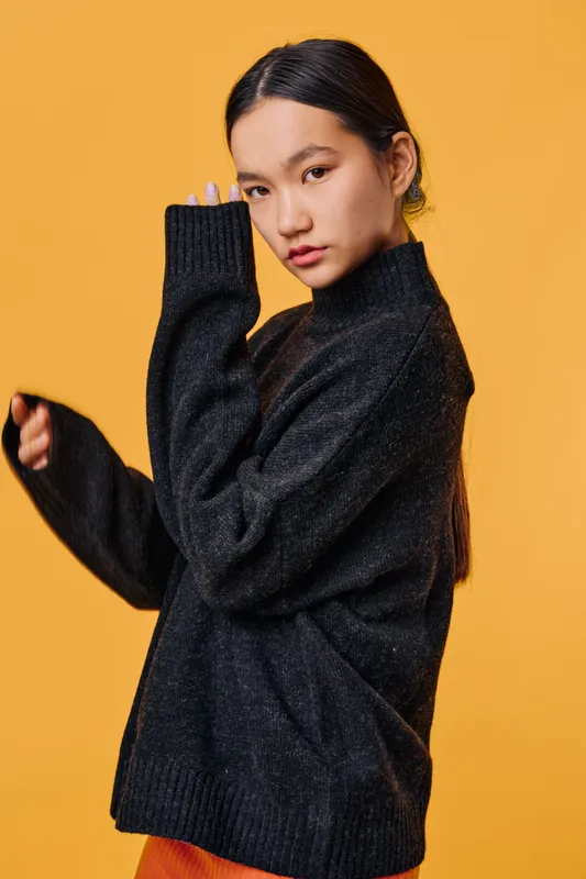 Young woman posing in black knitted sweater with long sleeves against orange background. Portrait of serious Asian female model with dark hair and casual fashion style.