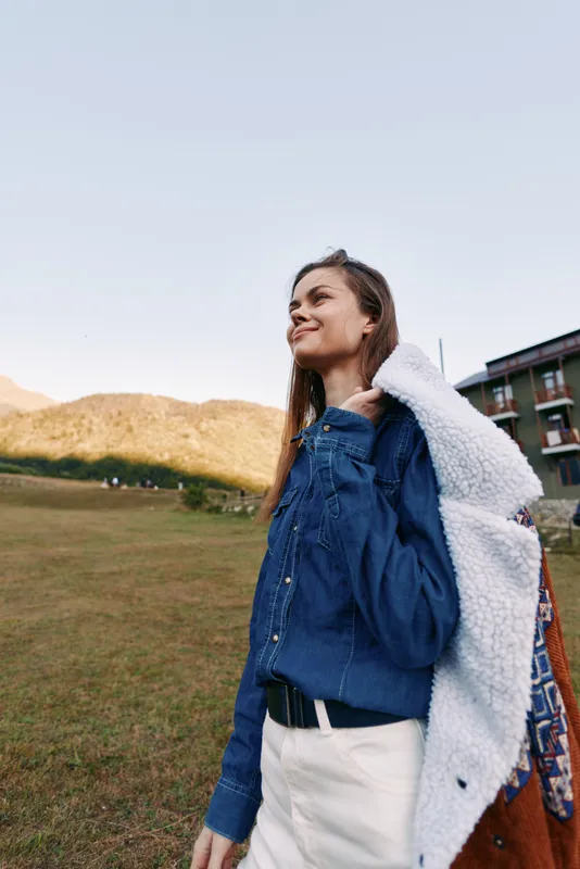 Woman smile jacket outdoors mountains field travel casual portrait lifestyle, young adult carrying a coat over her shoulder in grassy area near a lodge, happy confident and relaxed in nature.