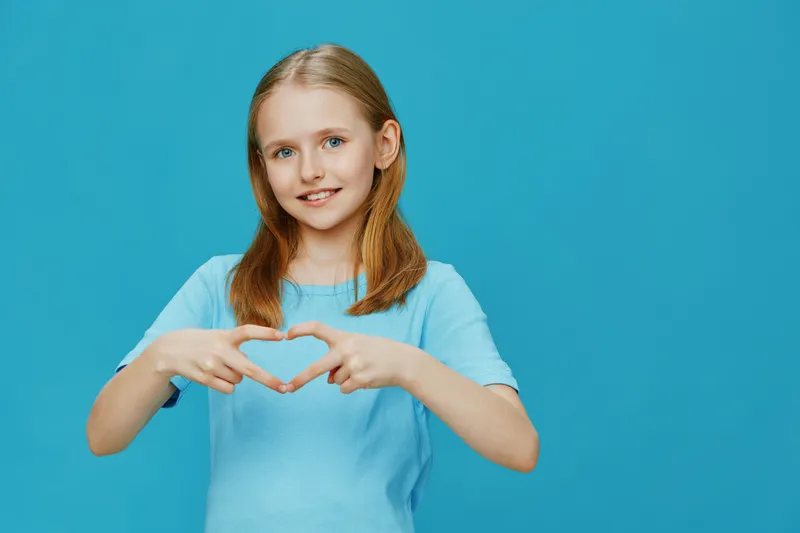 Cheerful young girl making a heart shape with her hands against a solid blue background, conveying love and positivity in a vibrant, fresh style