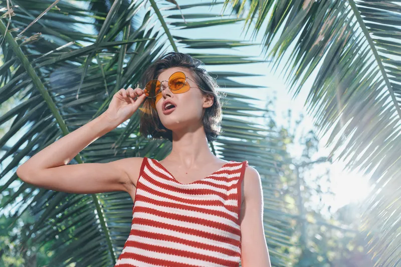 A stylish young woman wearing a red striped tank top and sunglasses stands under palm leaves in bright sunlight, enjoying a carefree moment outdoors.