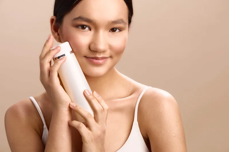 Asian woman gently holds a skincare bottle to her face, smiling softly in a clean studio. The scene conveys calm, care, and a confident beauty routine.