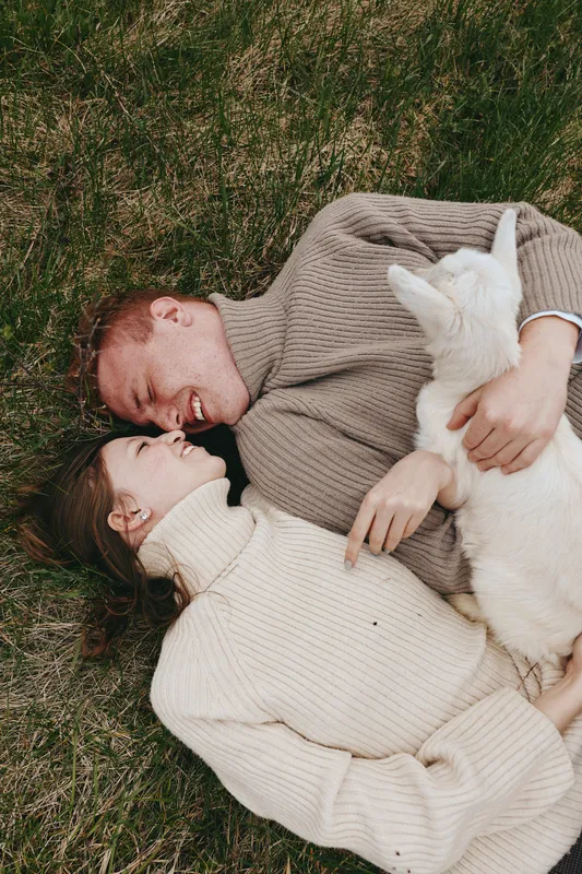 Loving couple resting outdoors with a goat kid, gentle smiles, cozy sweaters, sunlit grass, intimate moment, breeze, natural tones, countryside vibe, relaxed connection, candid lifestyle