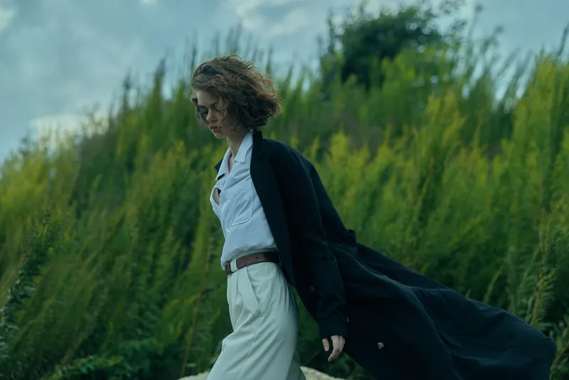 Woman in a white dress and flowing cloak walks through a windy field, showcasing fashion, movement, and natural scenery in a stylish outdoor moment.