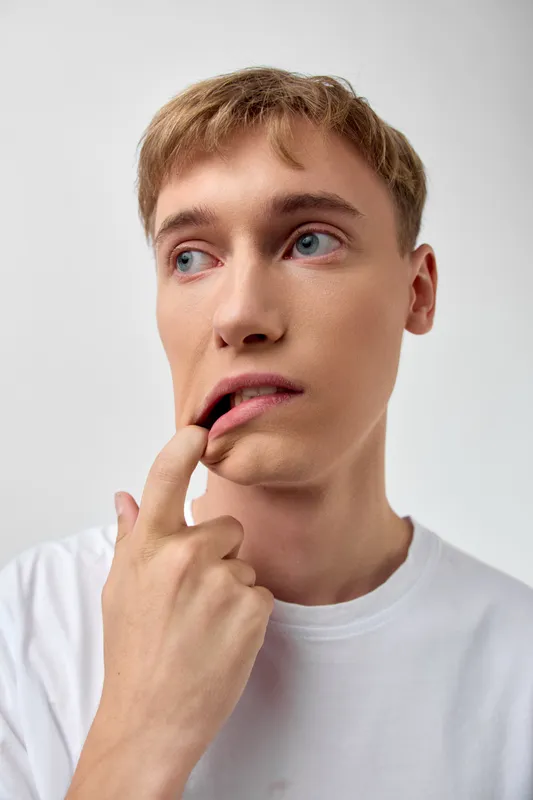 Thinking man touching lips in a white tee, pensive expression and close up mood, conveying uncertainty and contemplation in a clean studio with soft lighting, minimal background, and restrained