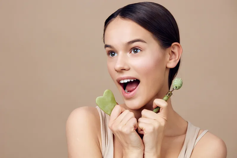 Young woman with a joyful expression holding skincare tools, representing healthy beauty and self care in a minimalist, neutral toned setting