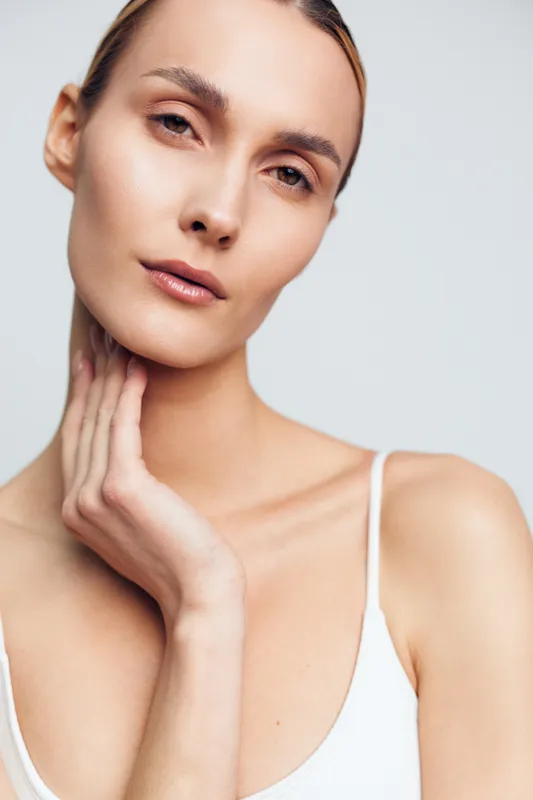 Young woman with clear skin gently touches neck showing natural beauty and calm expression in soft lighting against neutral background wearing white sleeveless top.