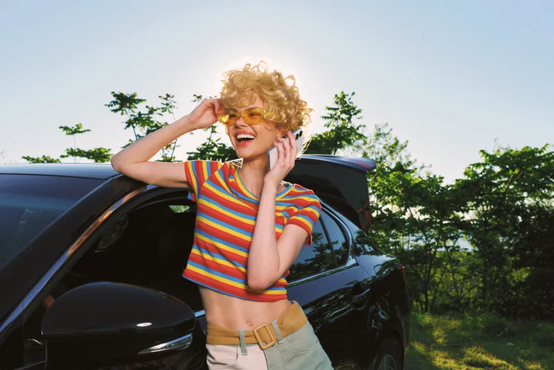 Joyful woman with curly blond hair in a striped crop top poses beside a car on a sunny day, showcasing casual lifestyle and retro film color ambiance. Bright outdoor portrait fashion cheerful summer