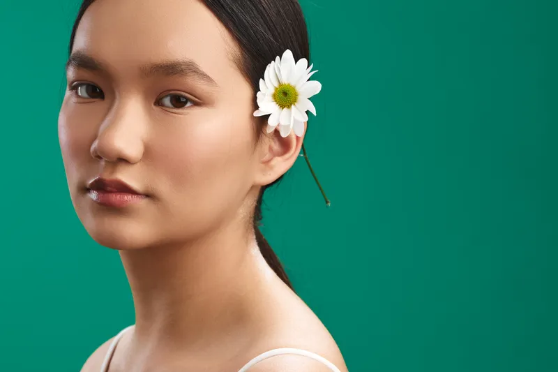 Asian woman portrait with a white daisy tucked behind her ear against a solid green backdrop, capturing calm confidence and natural beauty in a minimalist studio.