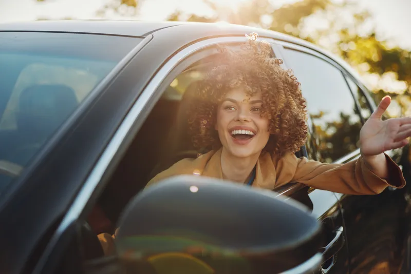 Joyful woman driver smiles from car window, capturing a lifestyle moment with retro film color, bright happiness, candid motion, sunlit street scene, cheerful ambiance.