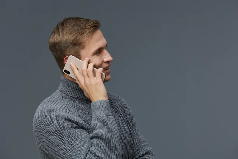 Modern young man with blonde hair wearing gray sweater talking on mobile phone isolated on plain gray background studio portrait. People lifestyle communication concept