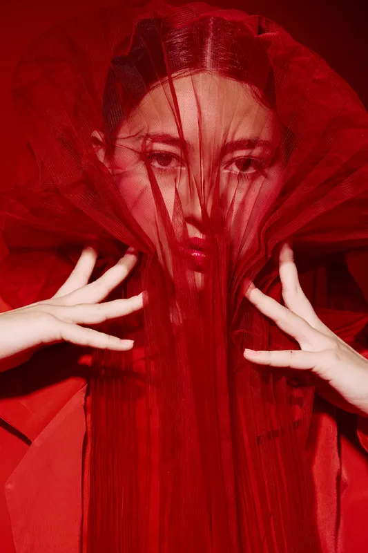 Female model with dramatic red tulle veil covering her face poses fashionably. Close-up beauty portrait with bold makeup and creative styling on red background.