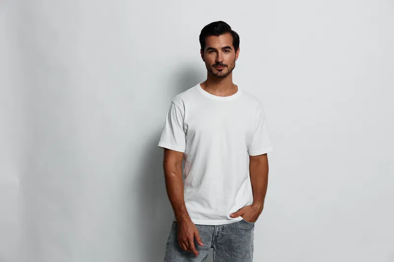 Confident young man wearing a casual white t-shirt poses with hands in pockets, smiling, against a plain light background in studio portrait style.