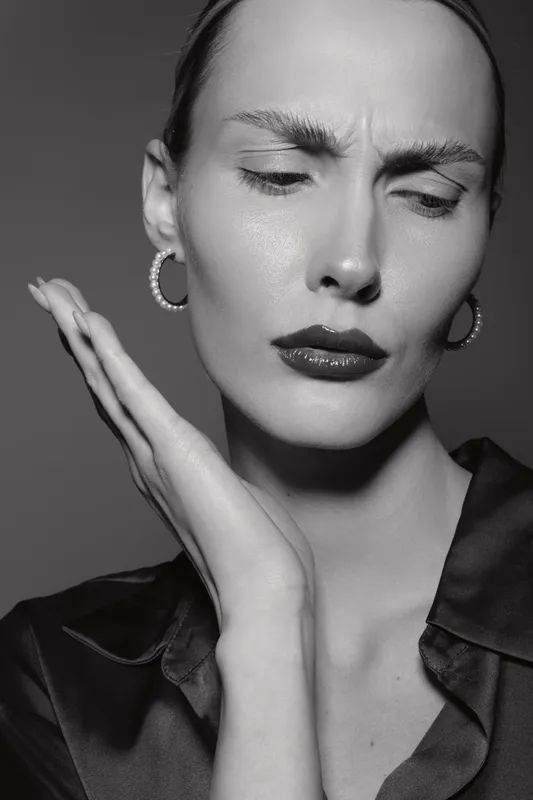 Caucasian woman with pale skin and slicked back hair shows discomfort holding face in black and white studio portrait.