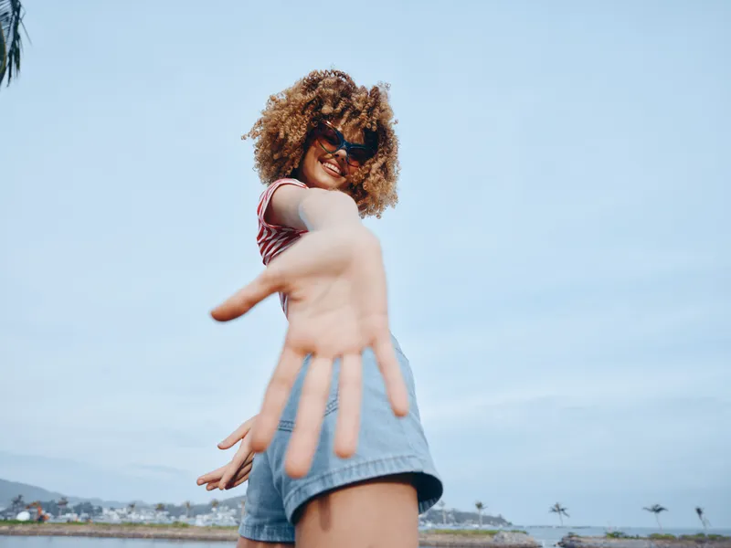 Vibrant beach portrait of a young person with curly hair, sunglasses, and denim shorts, reaching toward the camera with an energetic, joyful pose under a clear blue sky.
