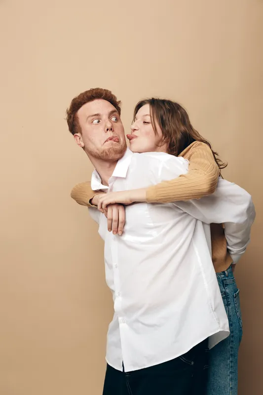 Young couple having fun with woman hugging man from behind on beige background. Casual clothing, playful mood, and affectionate moment captured in studio photography.