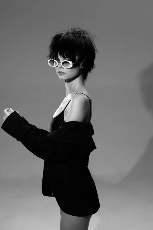 Black and white portrait of a stylish woman wearing sunglasses and an off-shoulder jacket with edgy short hair.