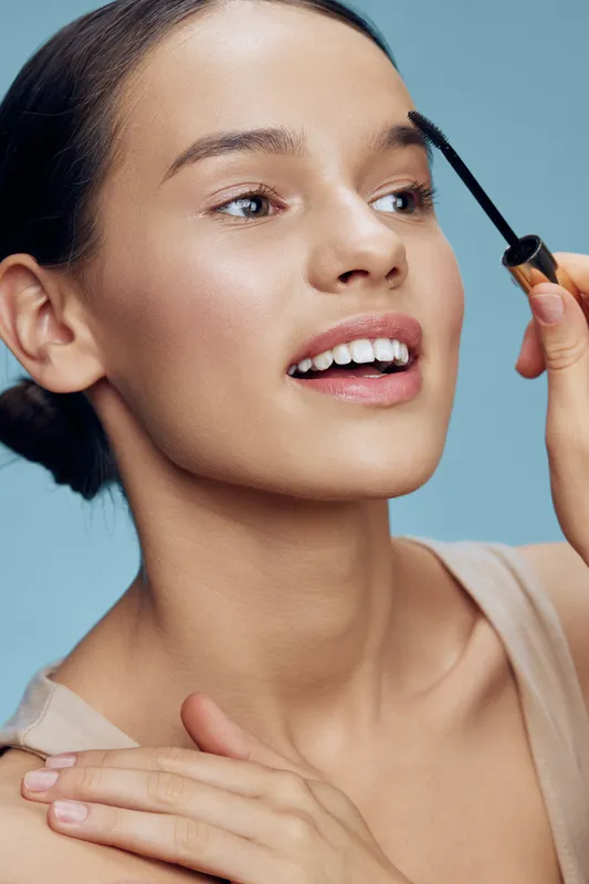Young woman applying eyebrow gel, showcasing a natural makeup look against a soft blue background for beauty and skincare concepts