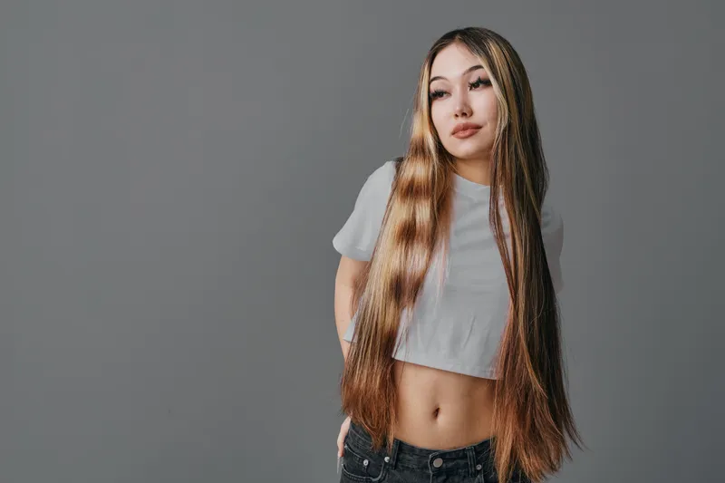 Young woman with long braided hair wearing grey cropped t shirt and black pants standing against plain grey background looking to side. Casual style and confident pose.