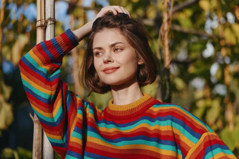 young woman, rainbow sweater, outdoor portrait, short brown hair, smiling woman and leaning on pole in sunny garden. 25-year-old model in casual outfit, relaxed lifestyle travel vibe.