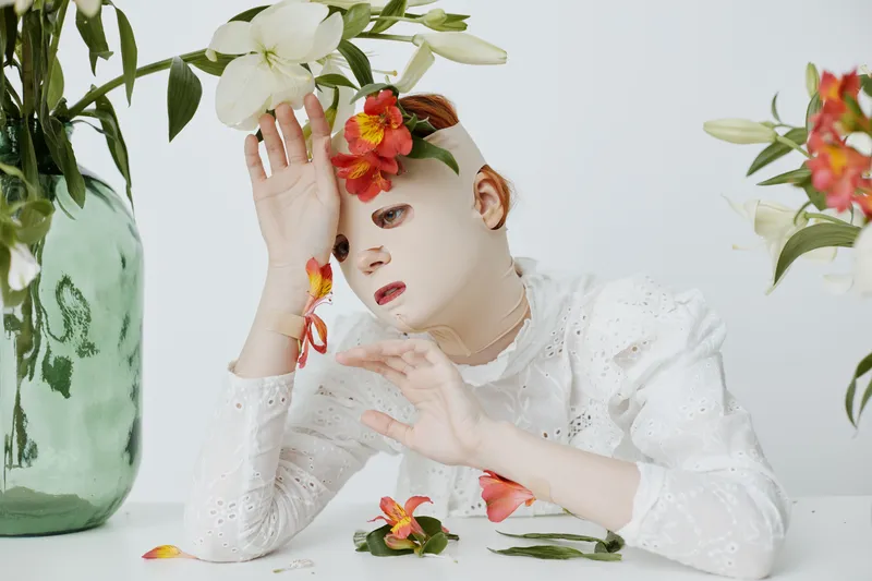 Artistic portrait of a model with floral elements, wearing a white lace dress, evoking emotions of tranquility and introspection in a minimalist setting