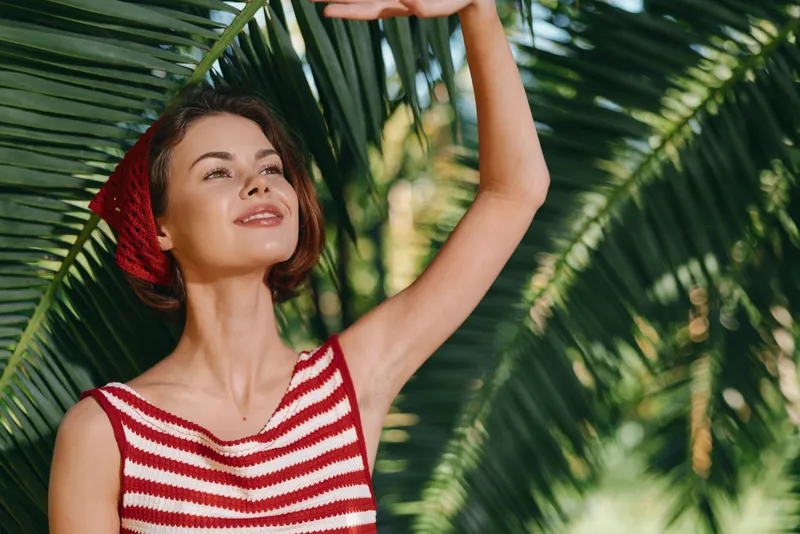 A cheerful woman wearing a striped red and white top stands among lush tropical leaves, smiling softly as she enjoys sunny outdoor warmth, radiating relaxed vacation vibes and carefree confidence.