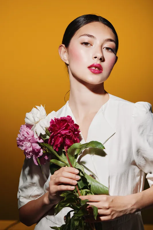 Young woman with slicked hair and glossy red lips holds vibrant peonies against warm yellow background. Creative beauty and fashion concept with elegant white dress.