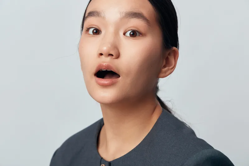 Young woman with surprised expression looking ahead, wearing dark clothing against plain light background. Emotional portrait with open mouth and wide eyes in studio lighting.