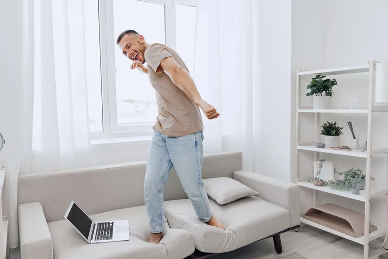 Happy man enjoying life while dancing in a modern living room. Bright, cozy interior with a laptop and plants creates a joyful atmosphere.