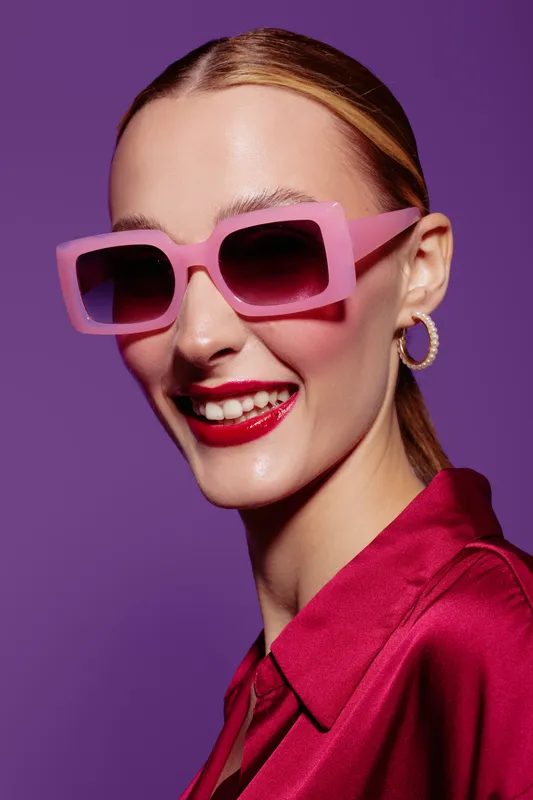 Smiling woman with pink sunglasses and red lipstick wears shiny red blouse against purple background. Fashion portrait with bright colors and confident expression for style concept.