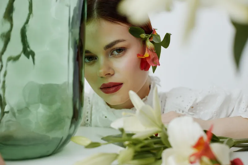 Beautiful young woman with floral accents near glass vase, evoking serenity and nature in a soft color palette, perfect for seasonal promotions and wellness themes