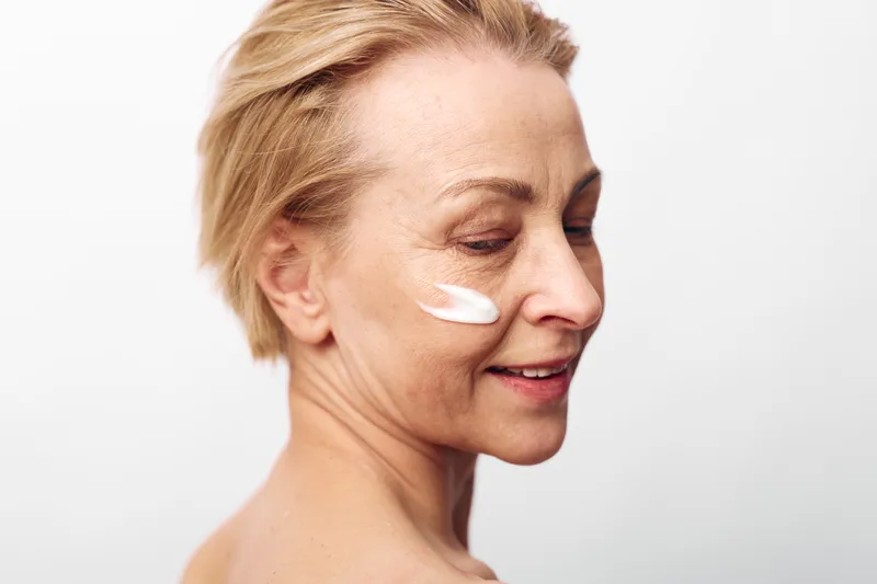 Mature woman with short blonde hair applies white face cream on cheek, smiling gently against white background. Concept of skincare, aging skin care, beauty routine, and self-care wellness.