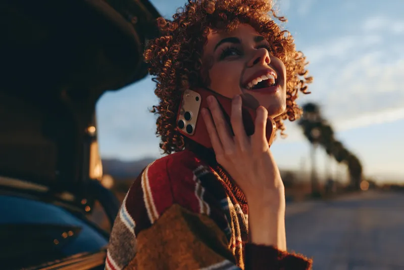 Woman lifestyle, smiling woman with curly hair talking on smartphone outdoors near car on road, warm evening light, candid communication moment, shallow depth, soft bokeh background, portrait