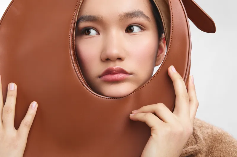 asian woman wearing a brown leather face mask accessory, posing in a studio with soft lighting. the unique prop frames her features, creating a bold fashion statement.