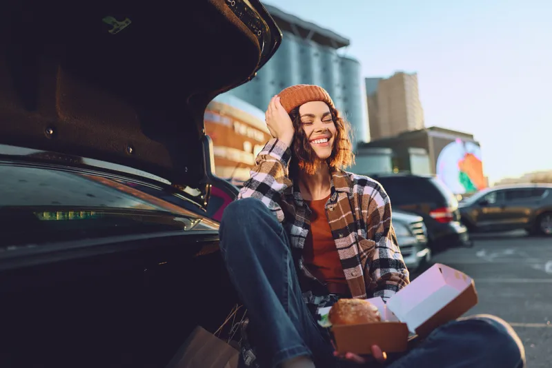 Woman eating food by car trunk with smile in urban parking, candid golden hour glow and authenticity. Cozy beanie, takeout meal and mindful living captured in emotional storytelling.