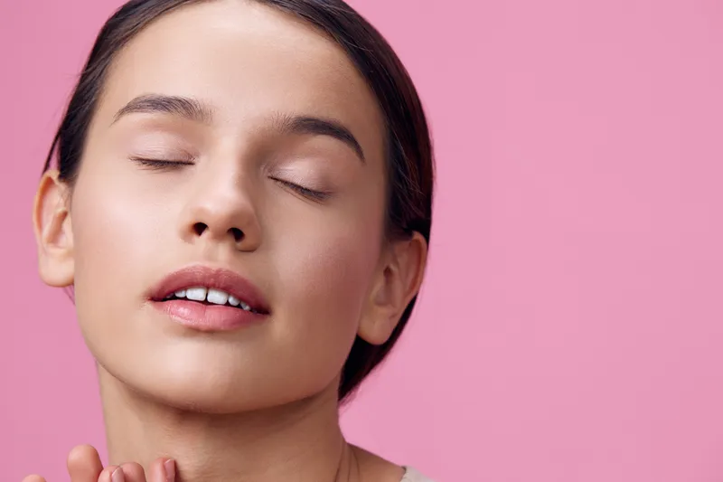 Young woman with closed eyes expressing serenity and bliss against a soft pink background, showcasing natural beauty and minimalistic style, ideal for wellness and self care themes
