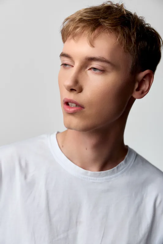 Man in a white shirt with a pensive expression, studio lighting highlights casual fashion, youthful vibe, calm gaze, modern elegance and a minimal background with subtle cotton tee