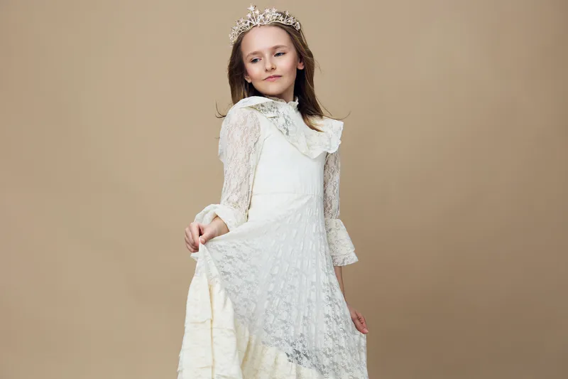 A young girl in a beautiful white dress with lace and a crown, exuding confidence and grace against a soft beige background, perfect for children s fashion photography