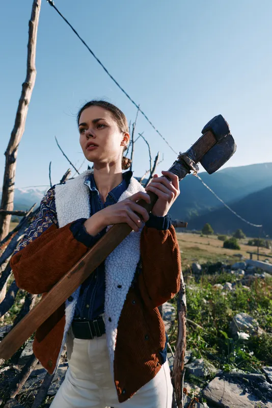 Woman with shovel on countryside farm near mountains, outdoor farming scene with agricultural tool and rural gardener in jacket preparing soil for planting or vineyard maintenance.