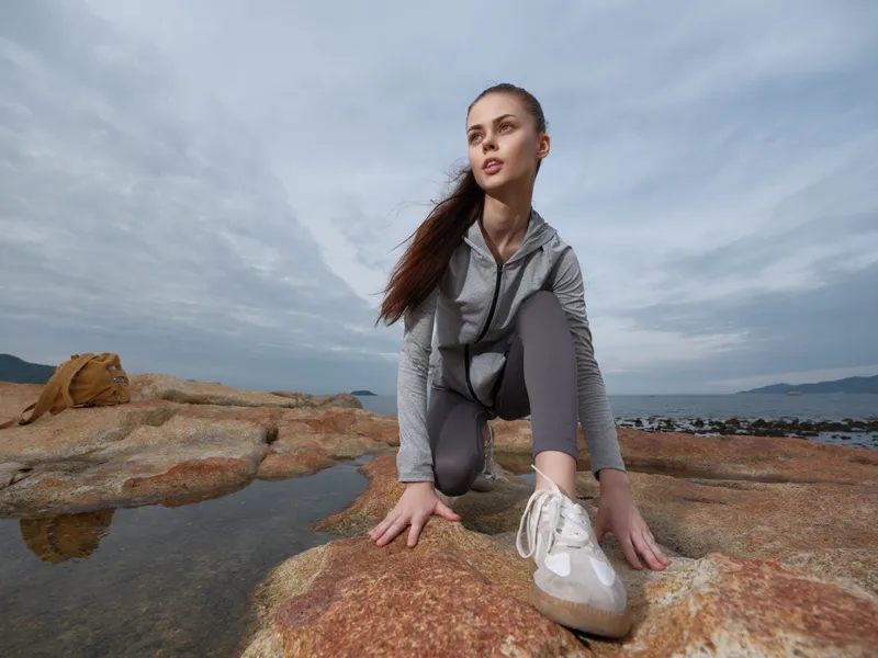 A focused young woman in athletic gear practicing yoga on rocky terrain by the sea, showcasing determination and calmness in a serene outdoor environment