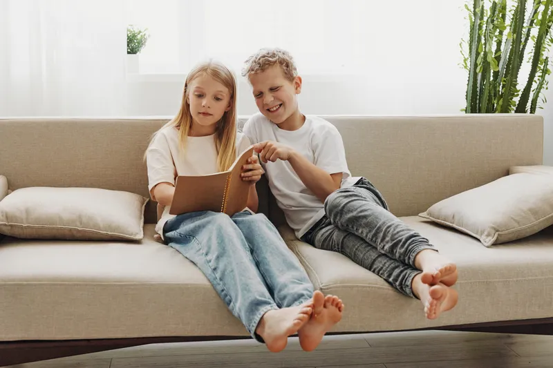 Two children sitting barefoot on a beige sofa reading a book together in a bright living room with plants. Concept of learning, childhood, and family bonding.