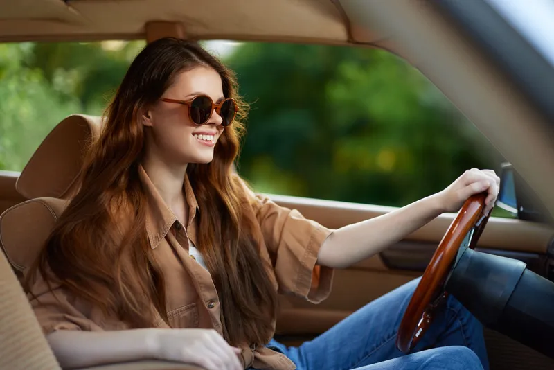 Young woman with long hair smiling while driving a car, wearing sunglasses and casual clothing, featuring a natural green background and a vibrant summer vibe.