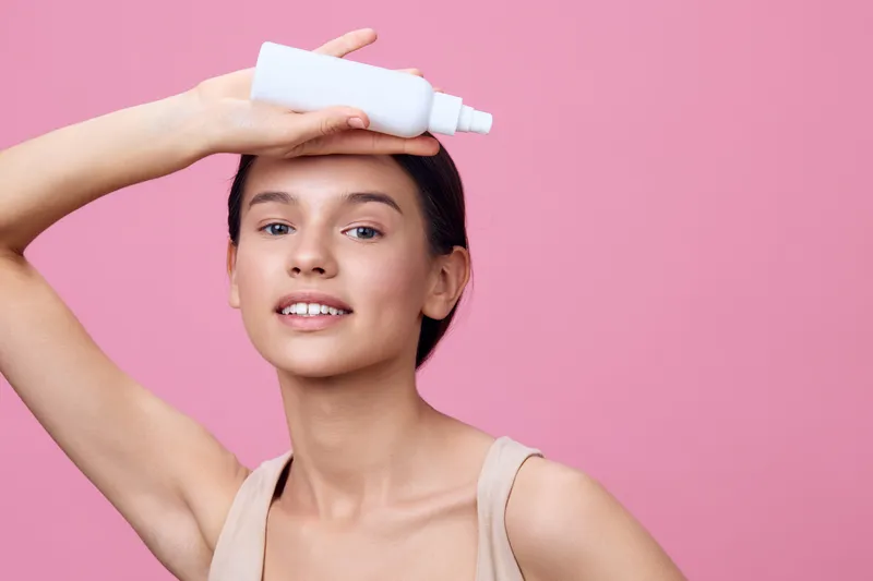 Young woman with clear skin applying skincare product on forehead against pink background, emphasizing beauty, confidence, and self care