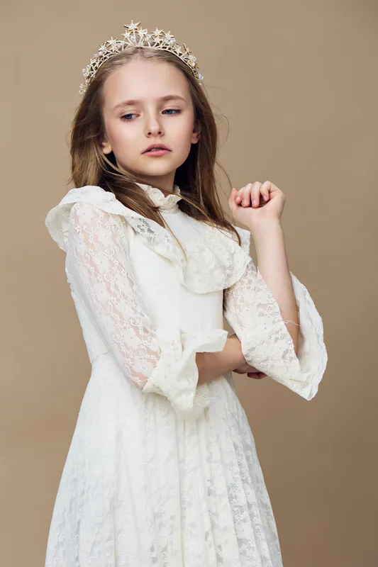 Elegant young girl in a lace dress with a crown, expressing a thoughtful and dreamy emotion against a neutral beige background, perfect for seasonal and fashion themes