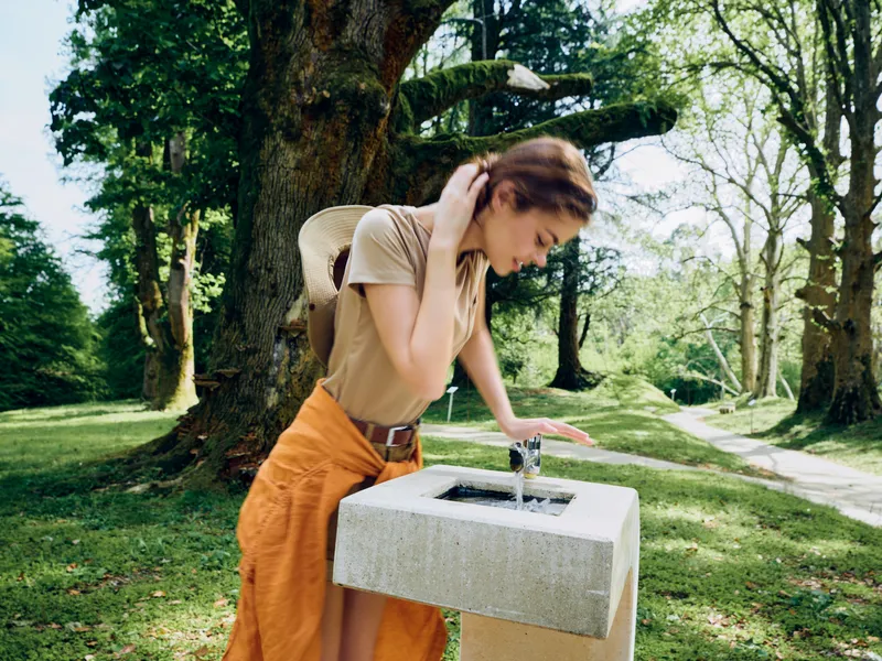 Woman at stone fountain in park, drinking and touching water with casual outfit and jacket tied around waist. Nature scene with tall trees, sunlight and peaceful outdoor moment