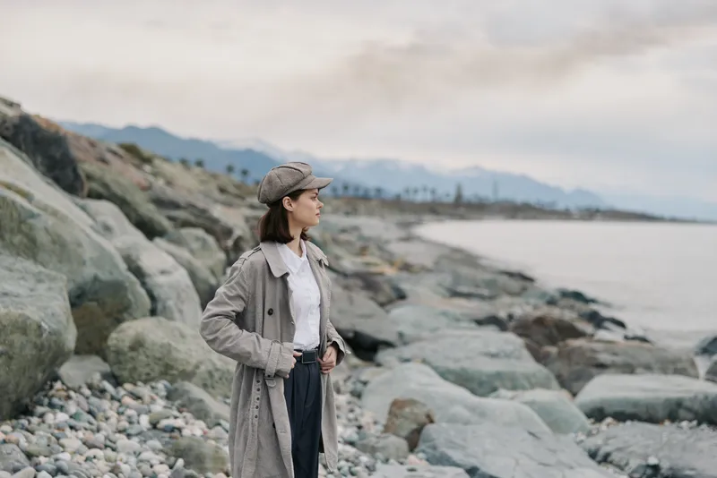 Young woman in cozy knitted cardigan and cap stands on rocky beach looking at sea. Cloudy sky and mountains in background create calm and contemplative atmosphere.