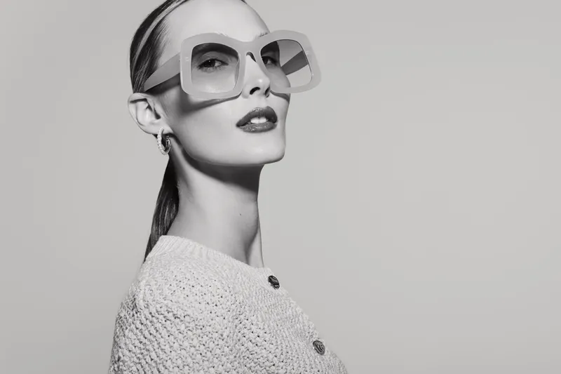 A stylish white woman with pale skin wears large geometric sunglasses and a textured sweater. The portrait in black and white highlights fashion and elegance concepts.