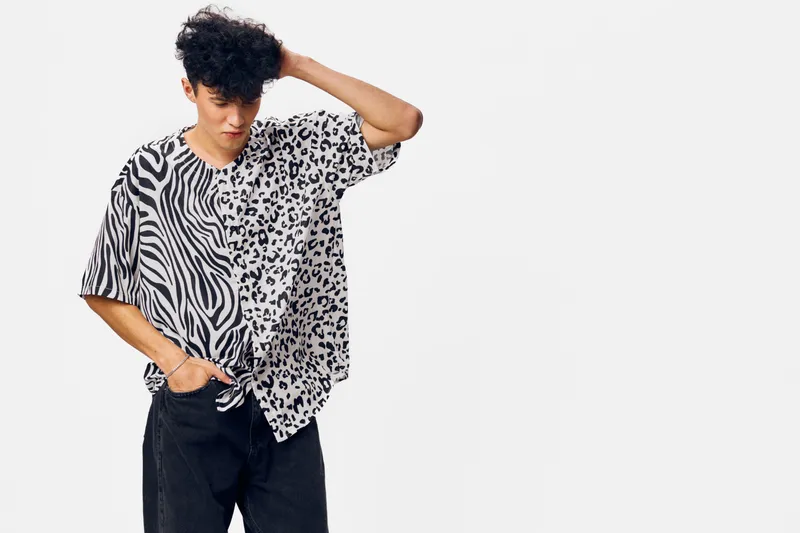 young man wearing mixed animal print shirt with zebra and leopard patterns looking down pensively against white background, casual style and modern fashion.